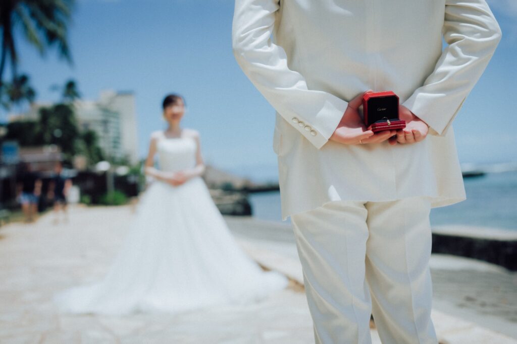 ハワイのビーチサイドで新郎が背中にプロポーズの結婚指輪ケースを持ち、奥にドレス姿の新婦が立つウェディングフォト